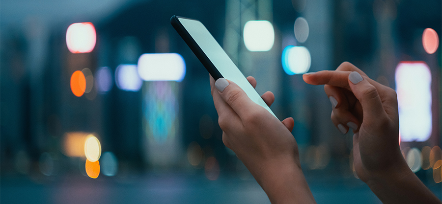 Close-up of hands using a smartphone at night, with blurred city lights and neon signs in the background.